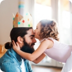 Father wears a hand-made paper crown, young daughter kisses his forehead.